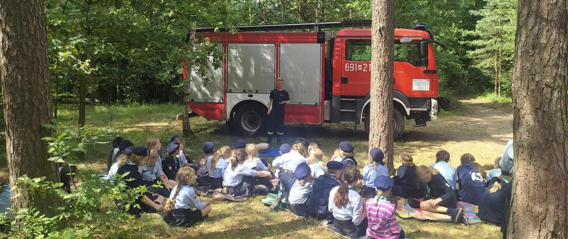 Strażak podczas spotkania z harcerzami. W tle samochód pożarniczy.