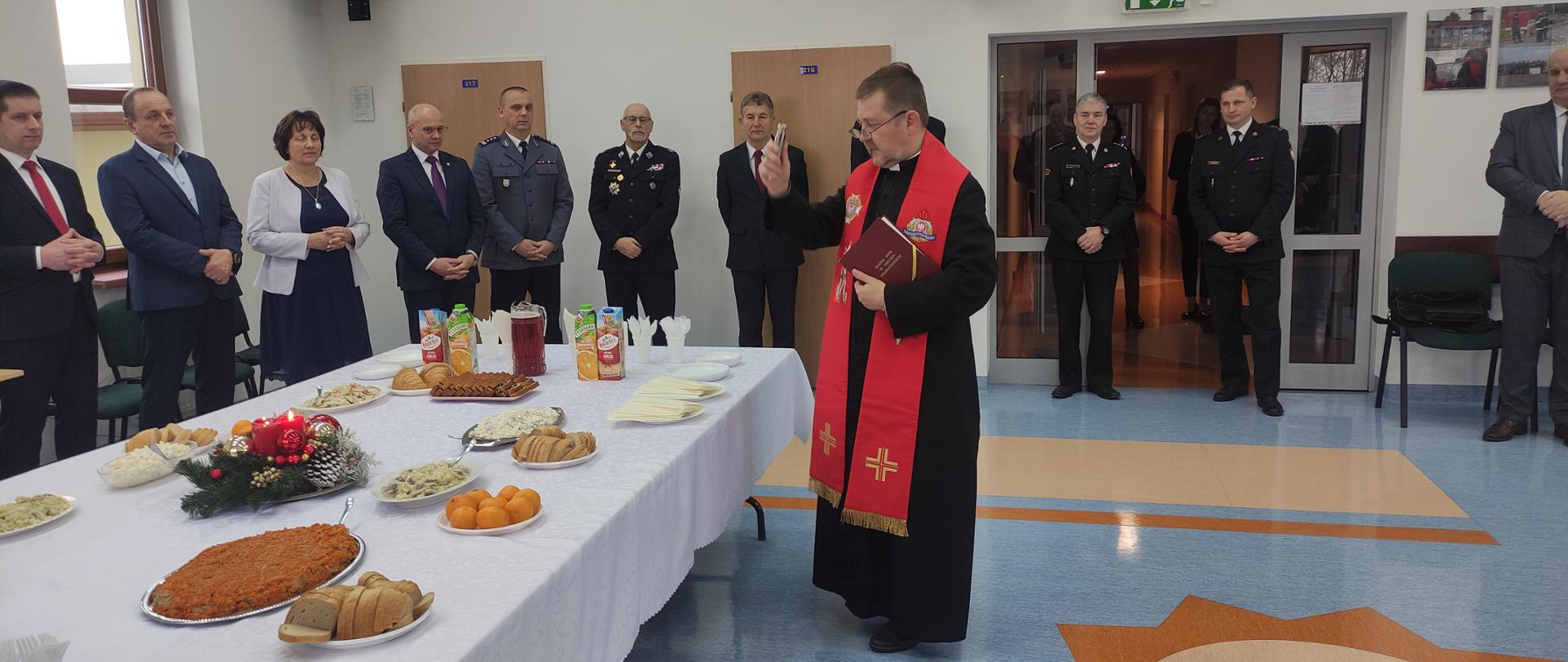 Spotkanie opłatkowe w Komendzie Powiatowej PSP w Sokołowie Podlaskim - W sali konferencyjnej komendy powiatowy kapelan strażaków ks. Krzysztof Kisielewicz święci pokarm i opłatek które są na stole. W tle pod ścianą stoją zaproszeni goście