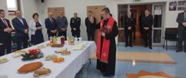 Spotkanie opłatkowe w Komendzie Powiatowej PSP w Sokołowie Podlaskim - W sali konferencyjnej komendy powiatowy kapelan strażaków ks. Krzysztof Kisielewicz święci pokarm i opłatek które są na stole. W tle pod ścianą stoją zaproszeni goście