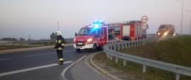 Zderzenie auta osobowego z motorowerem. Wiadukt nad drogą ekspresową S5. Na jezdni stoją dwa wozy straży pożarnej Pierwszy z nich ma włączone światła uprzywilejowania. Przed nim stoi strażak, który zabezpiecza miejsce zdarzenia. 
