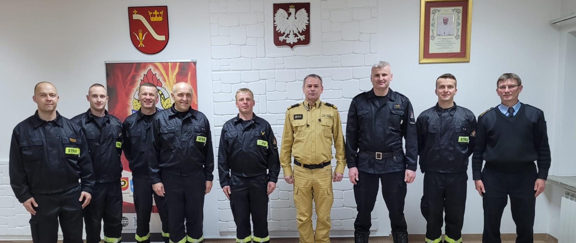 Strażacy w ubraniach koszarowych stoją w szeregu. W tle godło i herb powiatu. 