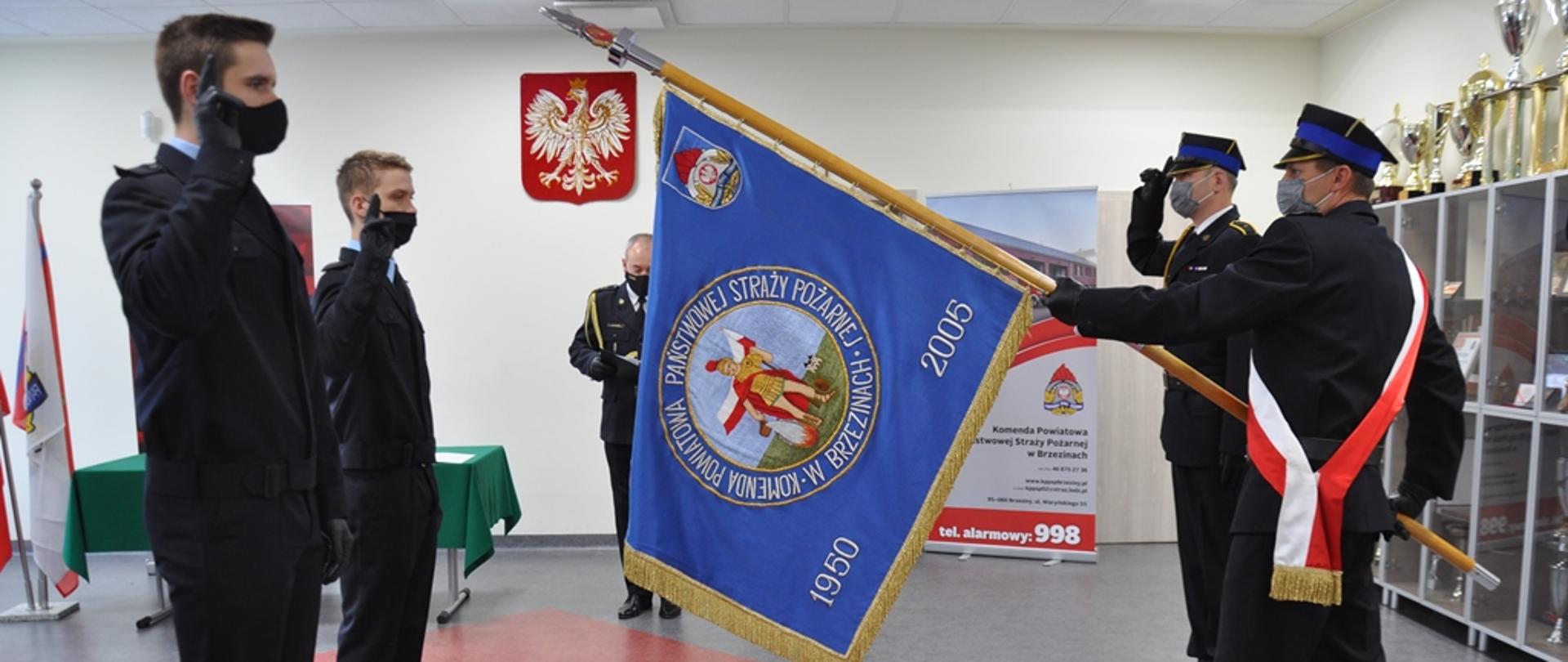 Poczet sztandarowy oraz dwóch nowoprzyjętych strażaków KP PSP w Brzezinach. w tle Komendant Powiatowy PSP w Brzezinach st. bryg. Dariusz Guzek. Zdjęcie w sali konferencyjnej komendy.