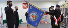 Poczet sztandarowy oraz dwóch nowoprzyjętych strażaków KP PSP w Brzezinach. w tle Komendant Powiatowy PSP w Brzezinach st. bryg. Dariusz Guzek. Zdjęcie w sali konferencyjnej komendy.