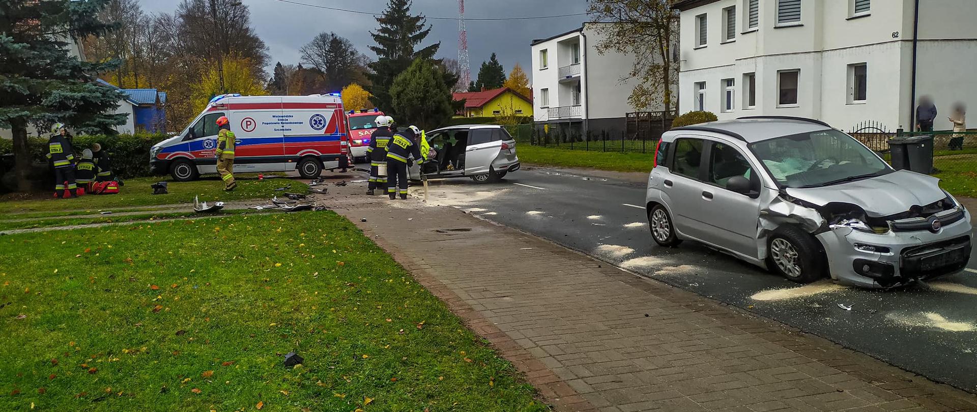Na zdjęciu znajdują się dwa rozbite pojazdu stojące na drodze , pierwszy z nich ma uszkodzony przód, drugi stoi bokiem na drodze ma pootwierane drzwi boczne, między nimi znajdują się strażacy którzy za pomocą sorbentu neutralizują płyny eksploatacyjne znajdujące się na chodniku oraz na jezdni. Z boku po lewej stronie na trawniku inni strażacy udzielają pomocy medycznej osobie poszkodowanej – obok stoi karetka pogotowia. W oddali widać samochód strażacki . Po obu stronach zdjęcia znajdują się zabudowania. Zdjęcie zrobione w porze dziennej, niebo pokryte jest chmurami.
