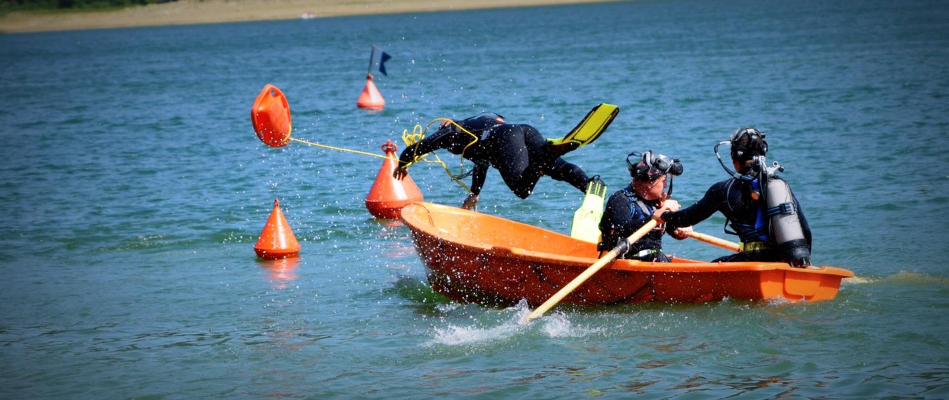 Zdjęcie zrobione nad jeziorem, podczas mistrzostw płetwonurków. Trzech strażaków nurków w łódce. Jeden z nich wyskakuje z łodzi do wody. 