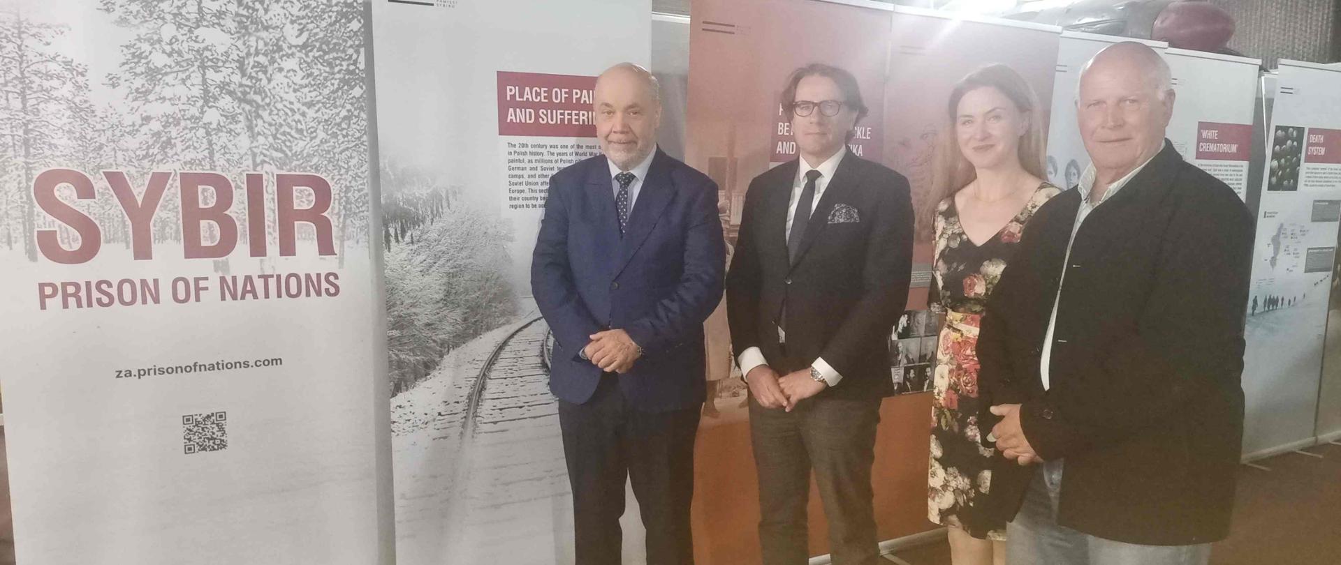 Grupa czterech osób pozuje do zdjęcia; od lewej strony charge d'affaires Jacek Chodorowicz, dyrektor Muzeum Pamięci Sybiru, prof. Wojciech Śleszyński, Julita Waś i prezes Związku Sybirakow Południowej afryki, Stefan Szewczuk; w tle plakaty wystawy Sybir. Prison of Nations
