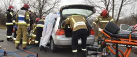 Zderzenie dwóch aut. Okolice Zaborowic. Odcinek jezdni, na którym doszło do wypadku. Strażacy wykonują dostęp do osób znajdujących się w volkswagenie stojącym na środku drogi. Przed samochodem znajduje się sprzęt hydrauliczny i nosze z karetki pogotowia. W tle drzewa.