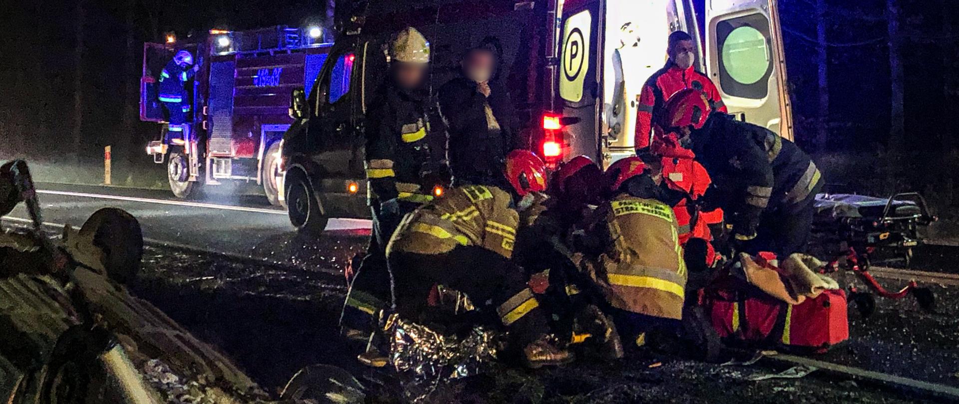 Na środku zdjęcia znajdują strażacy oraz ratownicy medyczni, którzy udzielają pomocy osobie poszkodowanej. Za nimi znajduje się karetka i wóz strażacki. W lewej części fotografii znajduje się samochód osobowy leżący na dachu w rowie. Pojazdy mają włączone światła błyskowe - jest noc.
