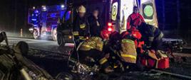 Na środku zdjęcia znajdują strażacy oraz ratownicy medyczni, którzy udzielają pomocy osobie poszkodowanej. Za nimi znajduje się karetka i wóz strażacki. W lewej części fotografii znajduje się samochód osobowy leżący na dachu w rowie. Pojazdy mają włączone światła błyskowe - jest noc.
