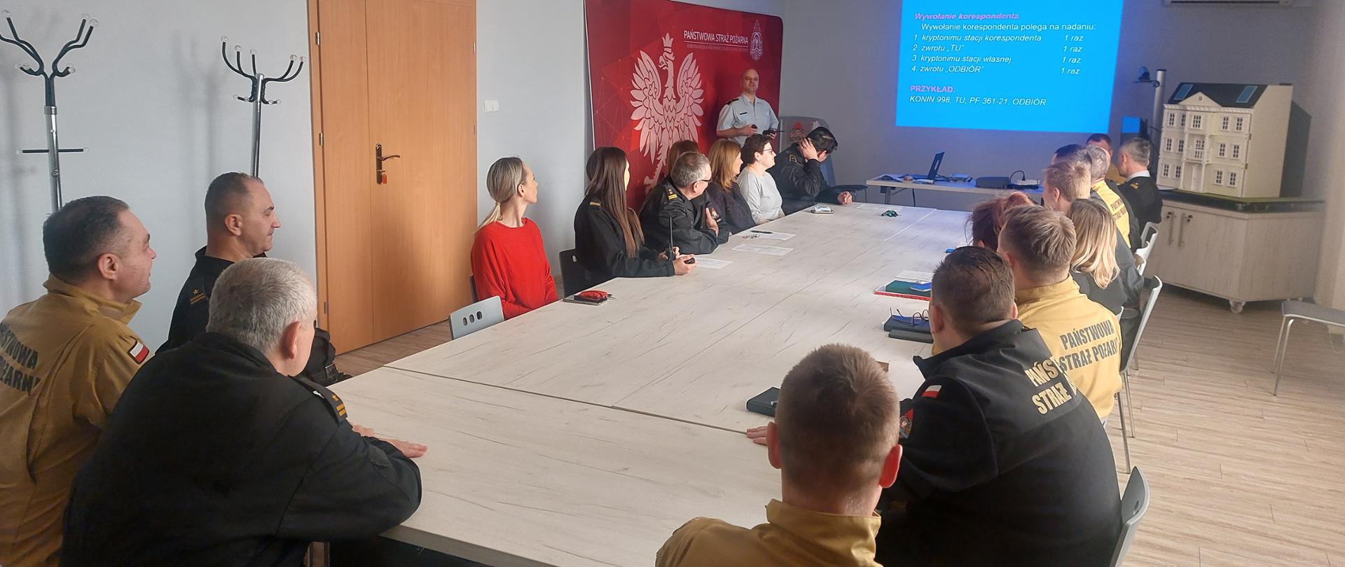 Zdjęcie przedstawia strażaków siedzących na świetlicy podczas szkolenia z łączności. Jeden ze strażaków za pomocą prezentacji multimedialnej omawia zagadnienia z łączności