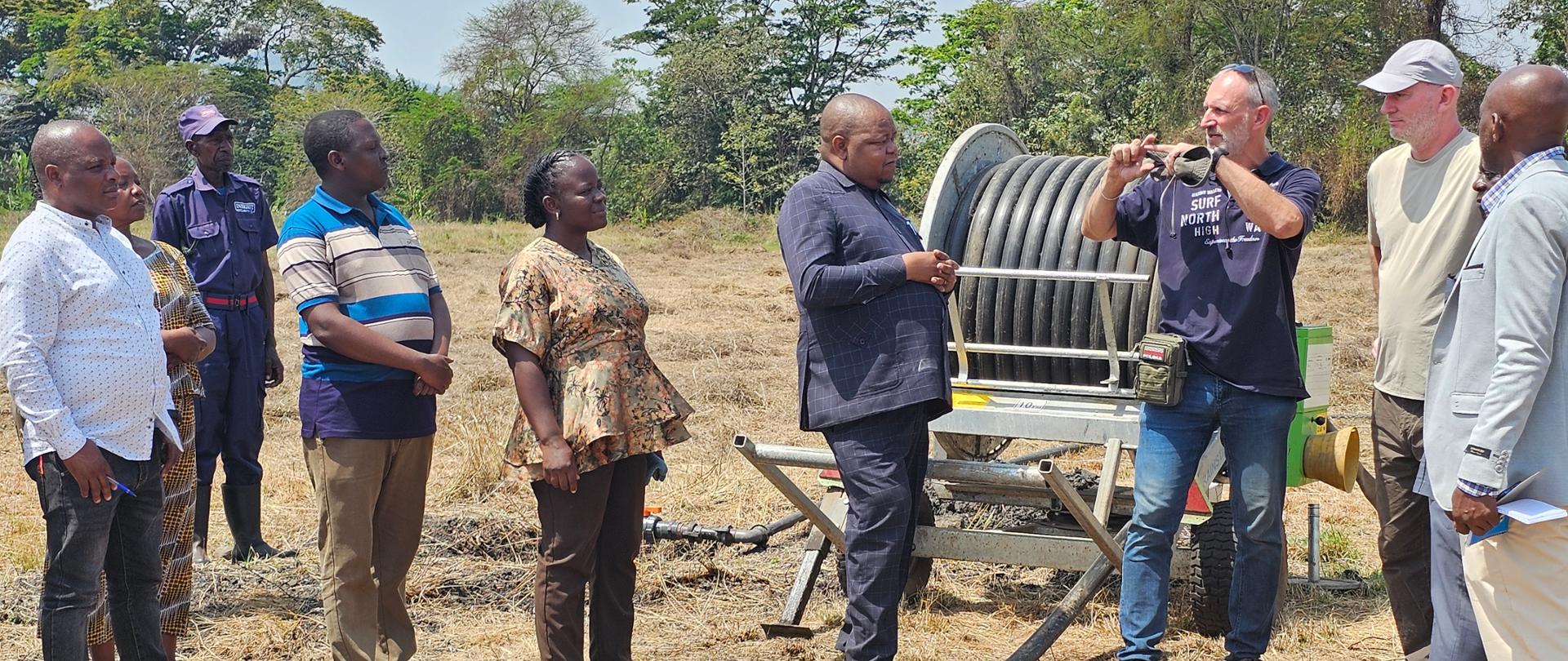 Mężczyzna coś opowiada i pokazuje rękoma, grupa osób mu się przygląda, stoją na wypalonej od słońca trawie, widać długi wąż ogrodowy