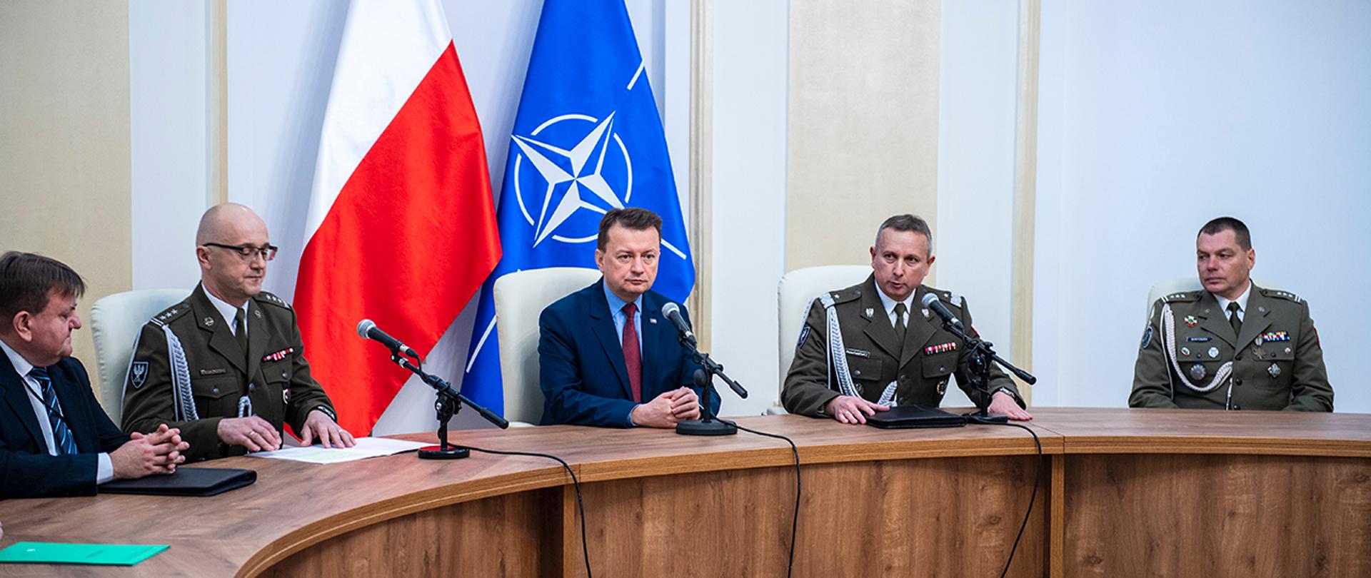 Szef MON siedzi przy stole z dowódcami Wojska Polskiego. Za nim flagi Polski i NATO.