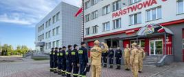 Strażacy w mundurach koloru czarnego i musztardowego stoją na tle budynku komendy oraz masztu flagowego z podniesioną flagą państwową