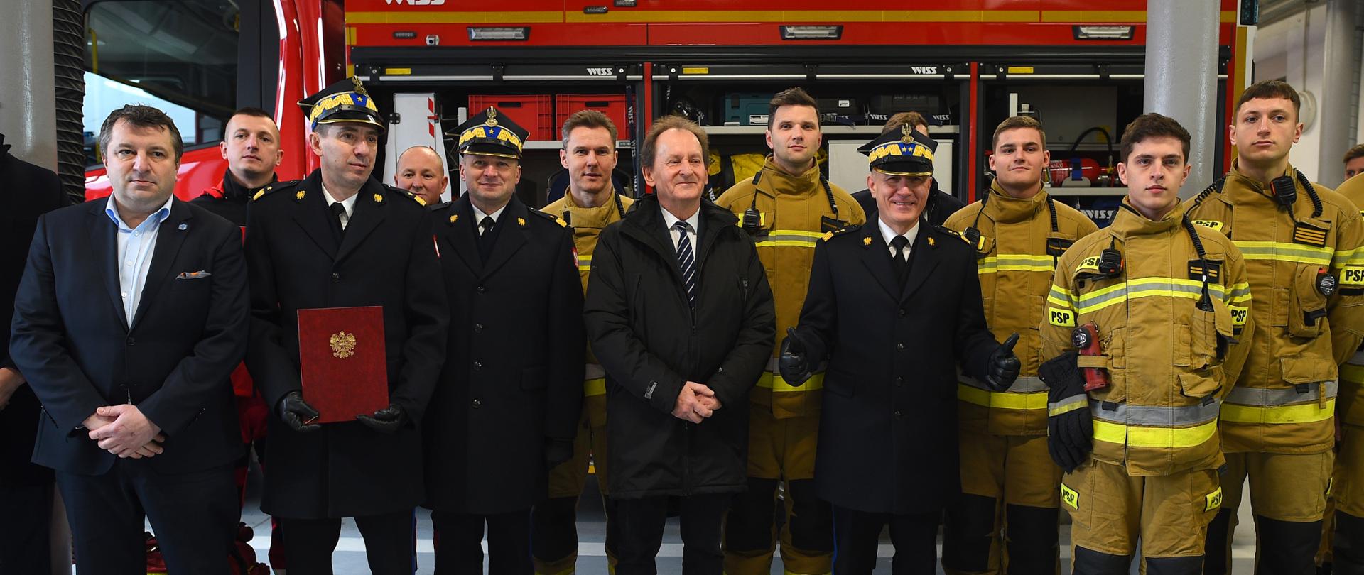 Wiceminister Bernacki stoi w dużej grupie ludzi, z lewej mężczyźni w garniturach i galowych mundurach, z prawej strażacy w żółtych ochronnych ubraniach, za nimi wóz bojowy z otwartymi lukami z wyposażeniem.
