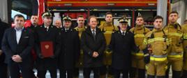 Wiceminister Bernacki stoi w dużej grupie ludzi, z lewej mężczyźni w garniturach i galowych mundurach, z prawej strażacy w żółtych ochronnych ubraniach, za nimi wóz bojowy z otwartymi lukami z wyposażeniem.
