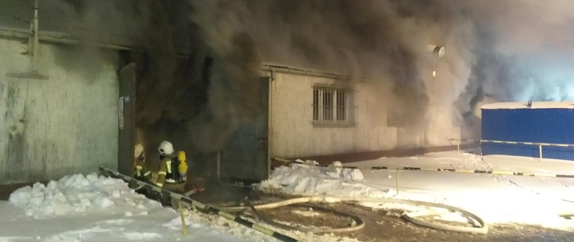 Na zdjęciu przedstawiony jest budynek hali magazynowo-produkcyjnej, z której wydobywa się gęsty, ciemny dym przez otwory okienne oraz otwarte drzwi garażowe. Przed drzwiami na kolanach klęczy dwóch strażaków w ubraniach specjalnych, z założonymi hełmami na głowę oraz zabezpieczonych w linię gaśniczą. Za nimi rozwinięta jest linia zasilająca składająca się z węża pożarniczego. Strażacy na plecach mają założone aparaty ochrony dróg oddechowych. Na zewnątrz, przed budynkiem zalega warstwa śniegu.