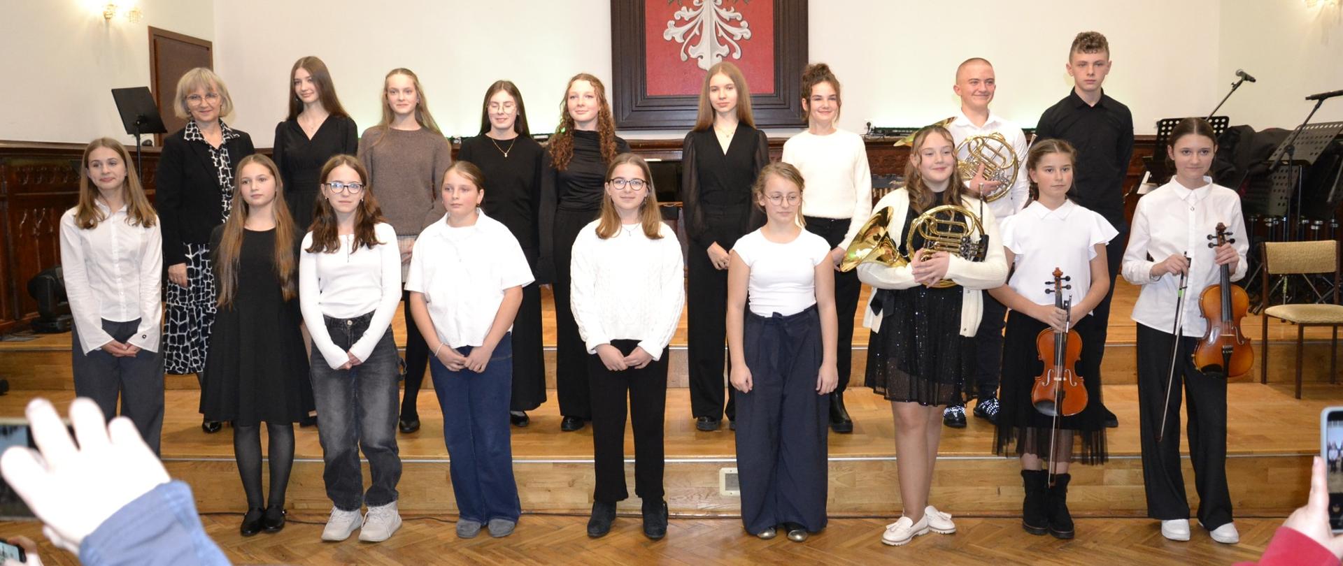 Grupa uczniów stojących na scenie Sali Królewskiej po koncercie klasowym; młodzi instrumentaliści z różnymi instrumentami oraz nauczycielka pozują do wspólnego zdjęcia na tle dużego godła z orłem.