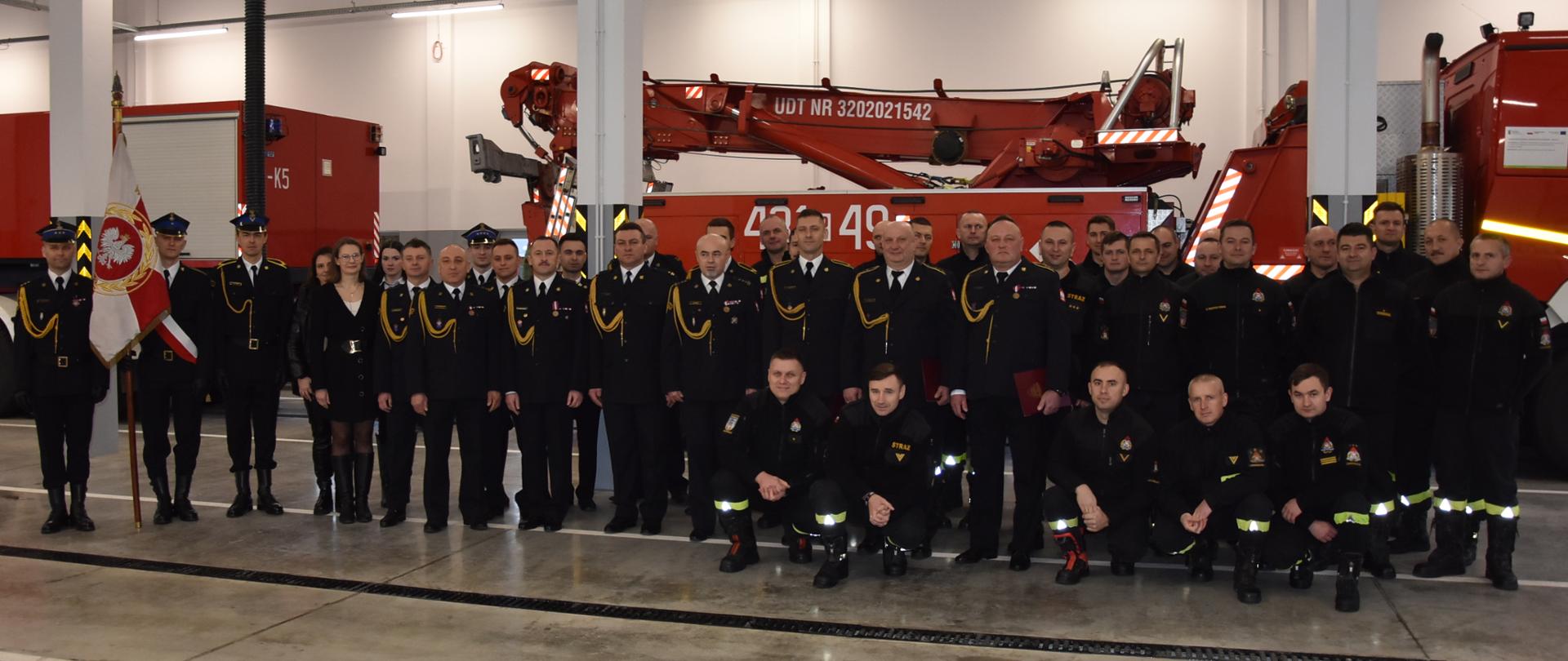 Zdjęcie grupowe wykonane w pomieszczeniach garażowych, przedstawiające zdjęcie strażaków w umundurowaniu wyjściowym i koszarowych. Po lewej stoi poczet sztandarowy.