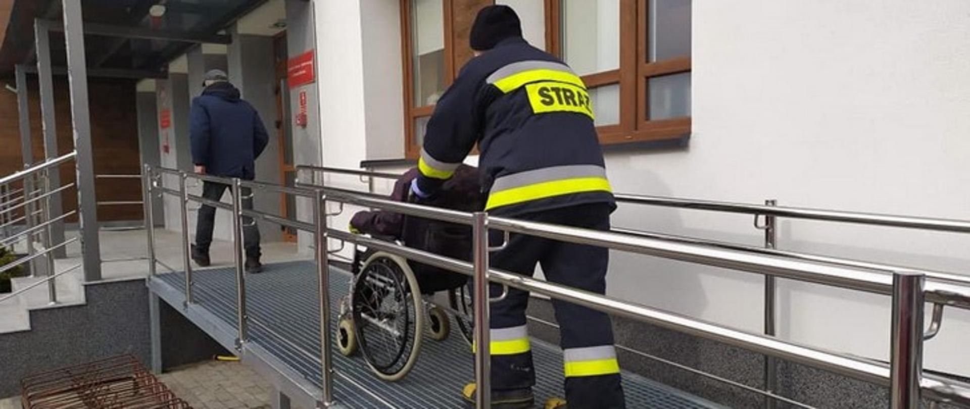 Zdjęcie przedstawia strażaka prowadzącego starszą osobę na wózku inwalidzkim.