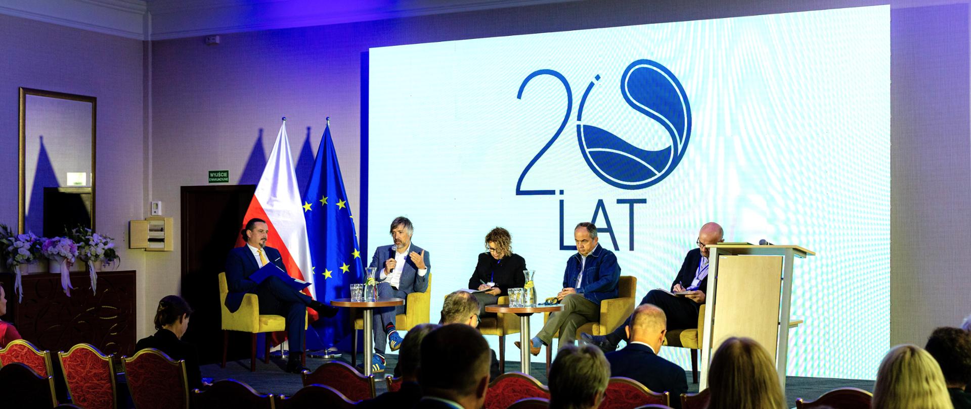 Panel dyskusyjny podczas konferencji. Na scenie pięć osób siedzi w fotelach i bierze udział w debacie, a przed sceną znajduje się publiczność. W tle widoczne są flagi Polski i Unii Europejskiej oraz napis „20 LAT”.