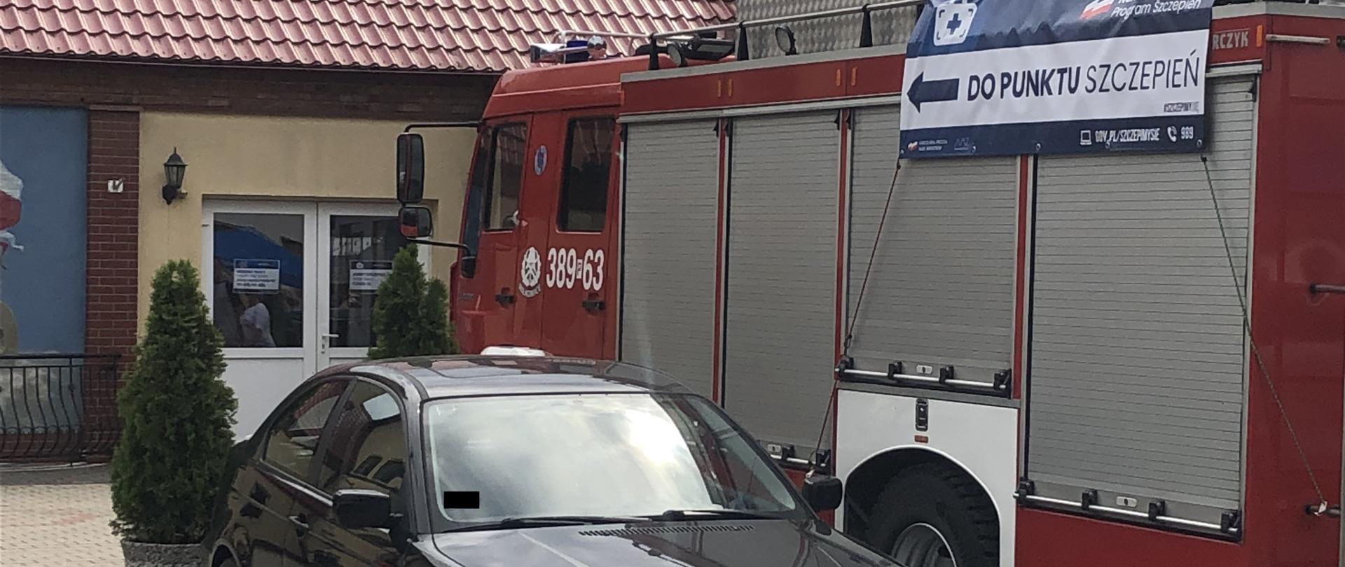 Samochód strażacki i samochód osobowy stoją na poboczu drogi przed budynkiem.