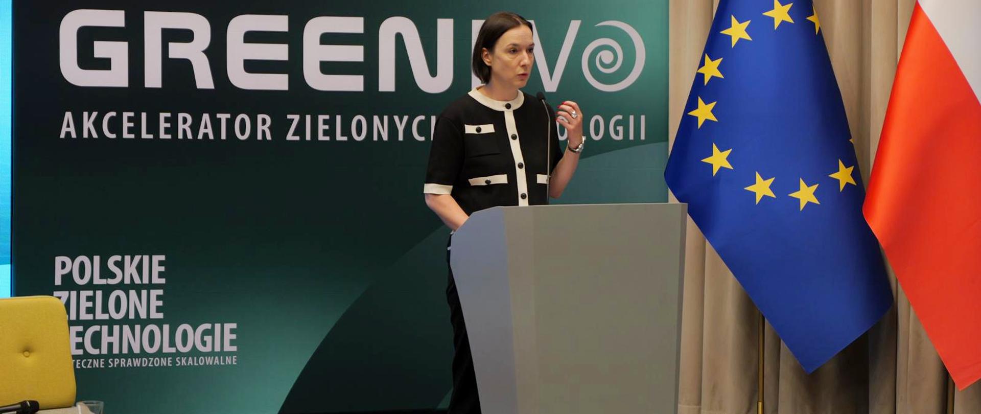 Wiceminister Eliza Zeidler stoi za mównicą. W tle ścianka i flagi. 