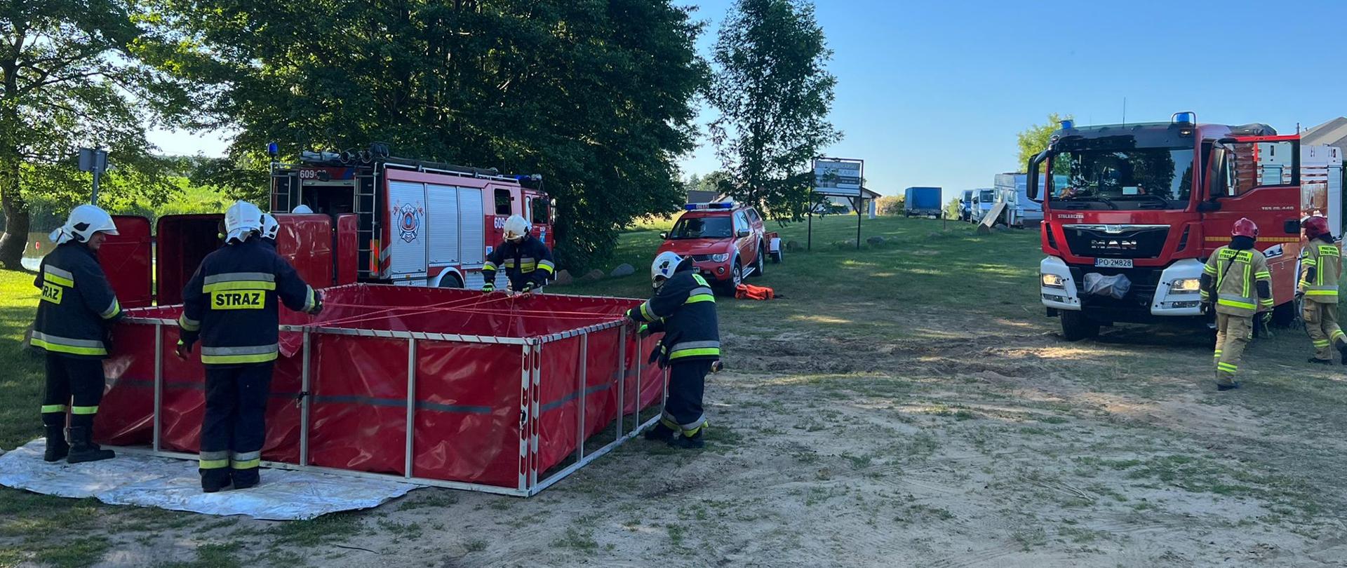 na zdjęciu strażacy podczas ćwiczeń samochody gaśnicze i specjalne na łące oraz strażacy budujący zbiornik wodny do celów gaśniczych
