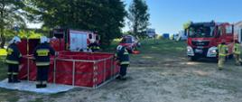 na zdjęciu strażacy podczas ćwiczeń samochody gaśnicze i specjalne na łące oraz strażacy budujący zbiornik wodny do celów gaśniczych