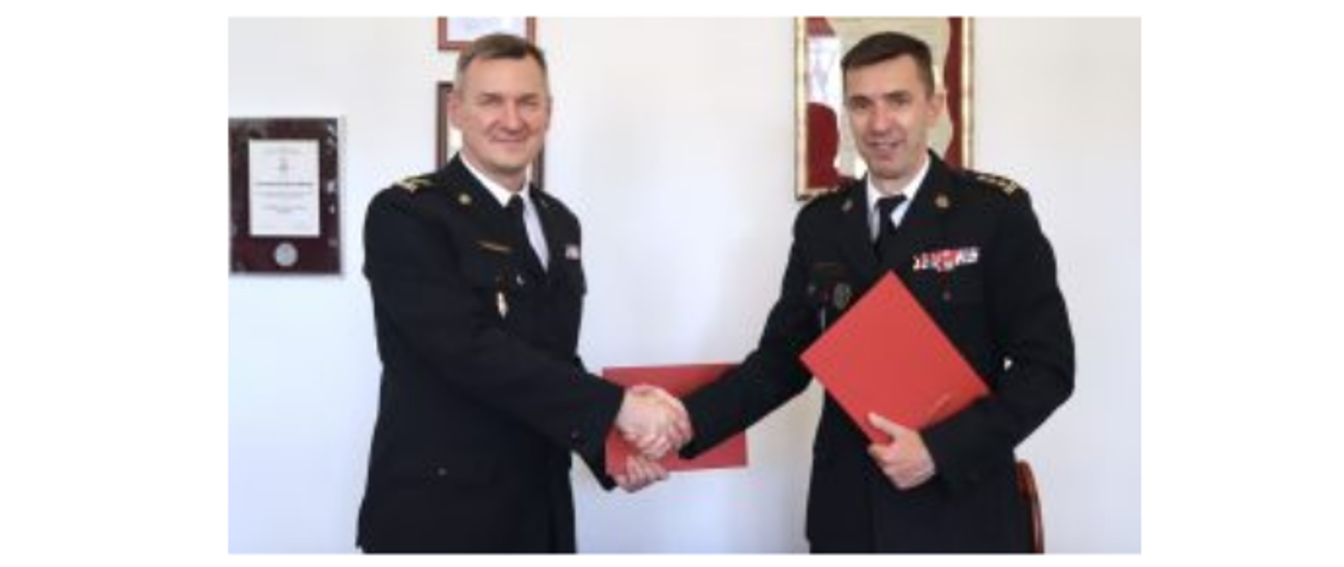 Starszy brygadier Leszek Smuniewski - Komendant Miejski Państwowej Straży Pożarnej miasta stołecznego Warszawy oraz starszy brygadier Mariusz Feltynowski - Rektor Komendant Szkoły Głównej Służby Pożarniczej podają sobie dłonie. W lewej dłoni obaj trzymają czerwone teczki. Ubrani są w mundury wyjściowe Państwowej Straży Pożarnej.