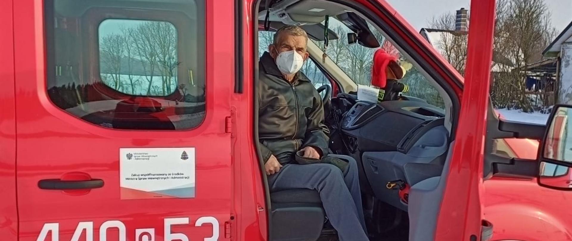Zdjęcie przedstawia kabinę czerwonego samochodu strażackiego widzianą z prawego boku. Na drzwiach biały napis 449c53. Przez otwarte drzwi widać ciemne wnętrze. Na fotelu pasażera siedzi mężczyzna ubrany w maseczkę, który zaczyna wysiadać. W tle widać leżący dookoła śnieg.