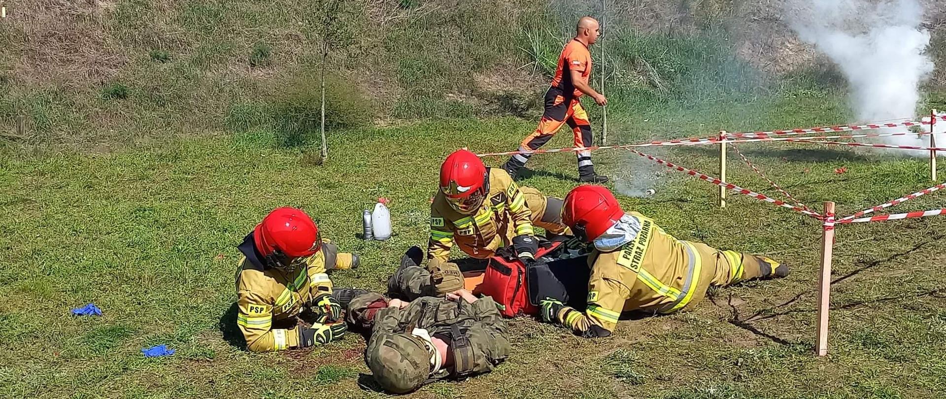 Manewry ratownicze. Teren byłego poligonu wojskowego w Sierakowie. Drużyna z Komendy Powiatowej PSP w Rawiczu realizuje zadania na piątym stanowisku. Strażacy udzielają kwalifikowanej pierwszej pomocy rannemu żołnierzowi. Trzech ratowników znajduje się wokół poszkodowanego w pozycji leżącej. Z prawej strony wbite w ziemię są drewniane paliki, pomiędzy którymi rozwinięta jest taśma ostrzegawcza. Strażacy maja na sobie ubrania specjalne koloru piaskowego i czerwone hełmy.