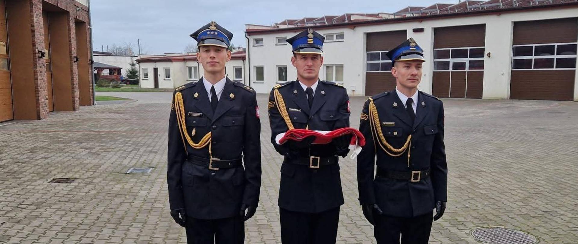 Na zdjęciu znajduje się poczet flagowy KP PSP Puck w składzie: kpt. Maciej Wesserling, st. sekc. Mateusz Gaffke oraz st. str. Wojciech Stachowicz. Funkcjonariusze ubrani są w mundury galowe PSP, z galowym sznurem na prawym ramieniu. Na głowach mają czapki rogatywki.