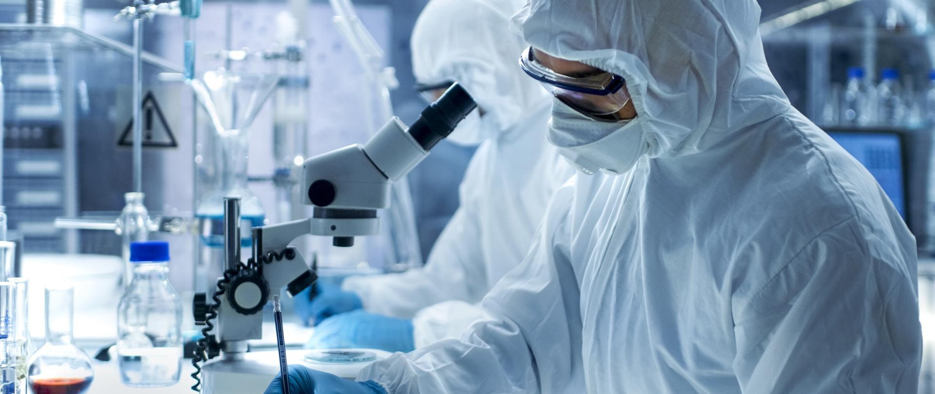 Dwie osoby w białych kitlach i nakryciach głowy podczas pracy przy mikroskopie
