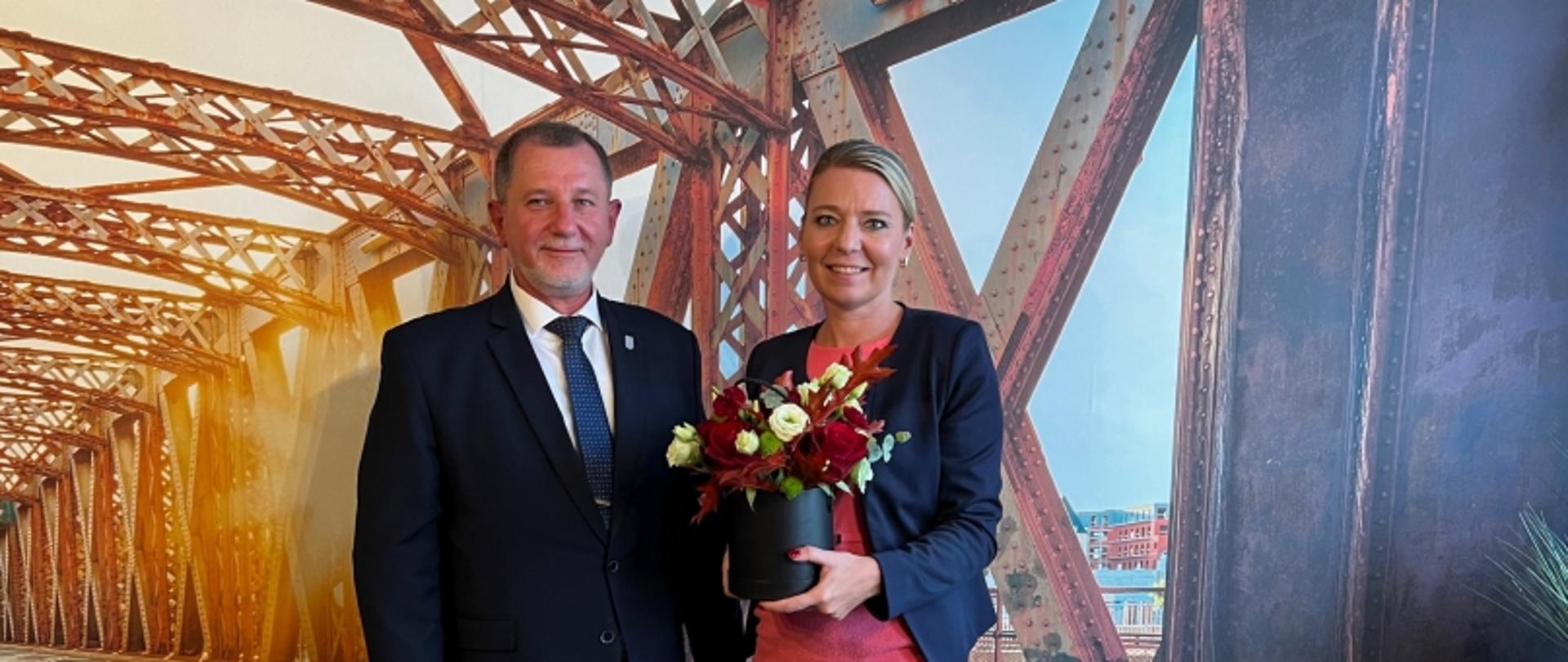 Dwie osoby stojące przed tłem przedstawiającym stalowy most z kratownicą, jedna z nich trzyma bukiet kwiatów.