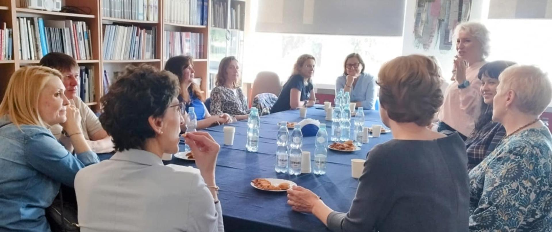 Zdjęcie ukazuje grupę osób – głównie kobiet – siedzących wokół dużego, prostokątnego stołu przykrytego ciemnogranatowym obrusem. Spotkanie odbywa się w jasnym, przestronnym pomieszczeniu, prawdopodobnie w bibliotece lub czytelni. Na stole stoją butelki z wodą i szklanki, a niektóre osoby trzymają w rękach kawałki ciasta lub przekąski.
W tle widać wysokie regały pełne książek oraz popiersia i obrazy zawieszone na ścianie. Na dużych oknach wiszą rolety z artystycznymi dekoracjami z tkanin. Jedna z kobiet stoi i mówi do pozostałych uczestników spotkania, które z uwagą jej słuchają. Panuje atmosfera skupienia i zaangażowania – wygląda na to, że jest to zebranie o charakterze edukacyjnym, kulturalnym lub dyskusyjnym.