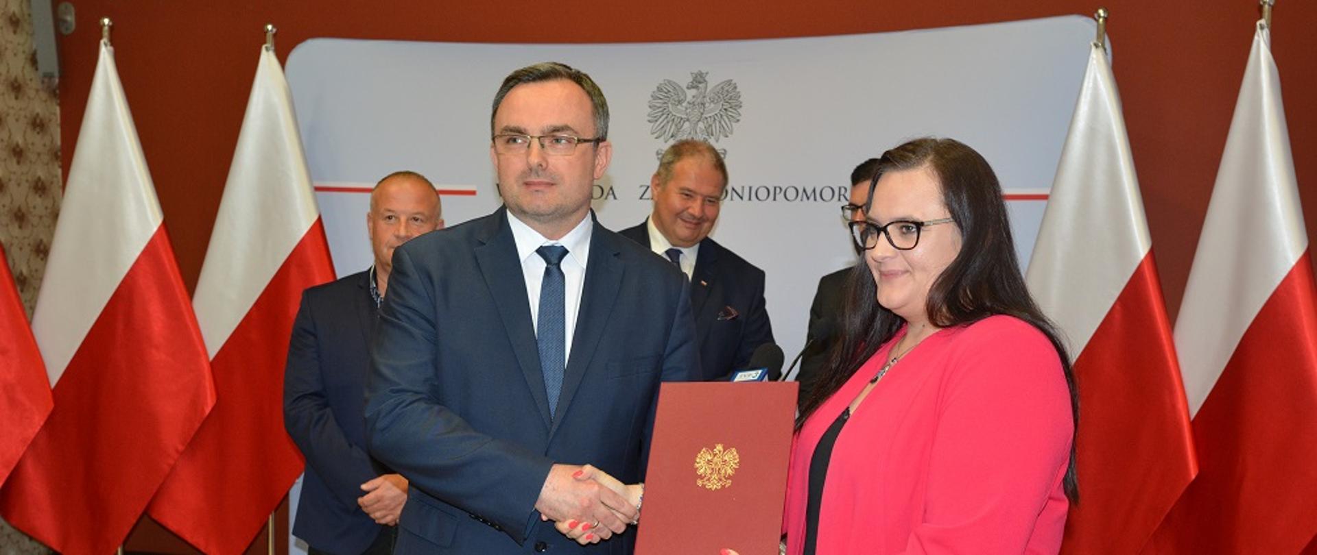 Na pierwszym planie minister Małgorzata Jarosińska-Jedynak ściska dłoń wojewody zachodniopomorskiego Tomasza Hinca. W tle stoi trzech mężczyzn przed flagami Polski
