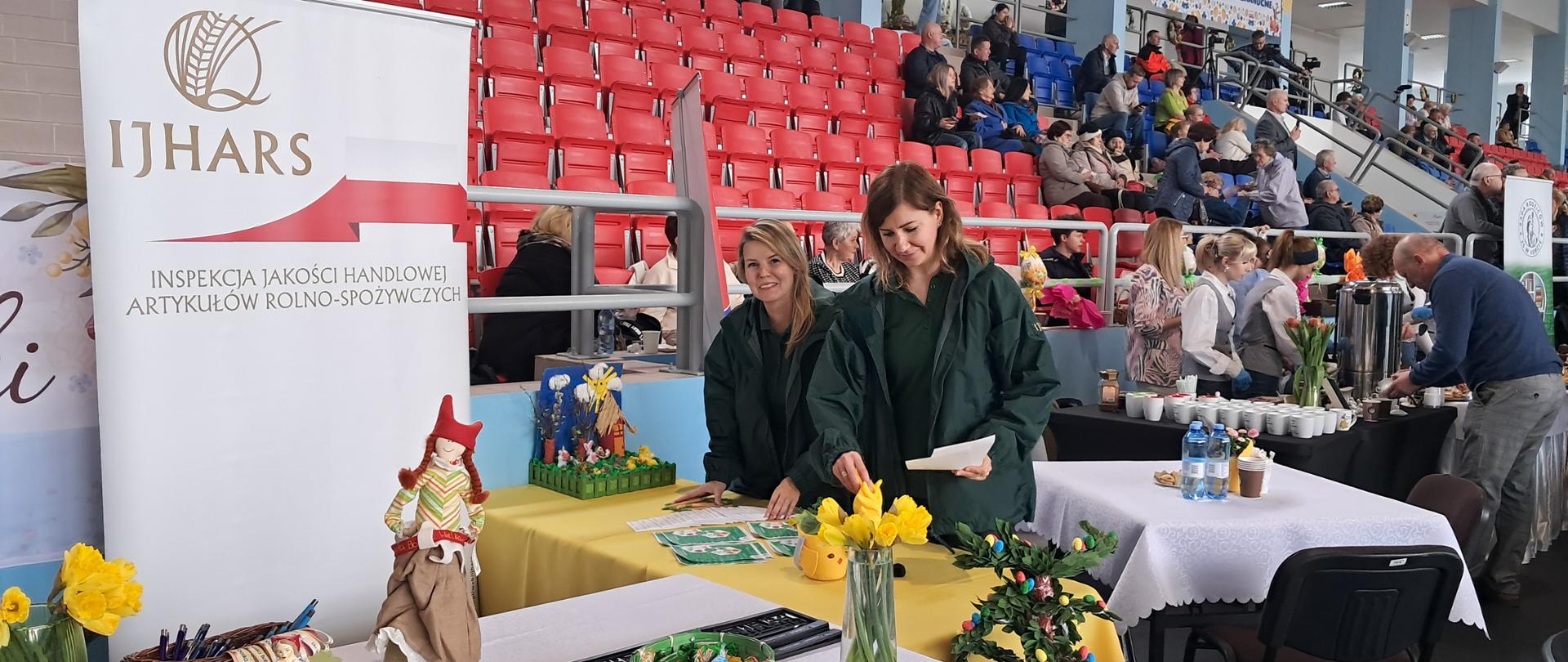 Stoisko IJHARS z dwiema inspektorkami stojącymi za wielkanocnym stołem. Przed nimi ustawione są świąteczne potrawy i dekoracje, a całość przykryta jest żółtymi obrusami. W tle widoczne jest logo IJHARS, podkreślające oficjalny charakter stoiska.