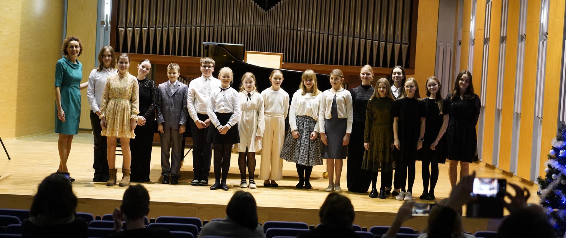 Zdjęcie w sali koncertowej. Na pierwszym planie publiczność, oraz puste fotele. Na scenie sali koncertowej w jednym rzędzie wszyscy uczestnicy koncertu. Z lewej strony nauczycielka prowadząca koncert. Za nimi fortepian, w tle organy. Z lewej strony przed sceną choinka bożonarodzeniowa udekorowana w łańcuchy i bombki.