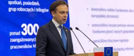 Minister Andrzej Domański stoi za mównicą z flagami Polski i Unii Europejskiej