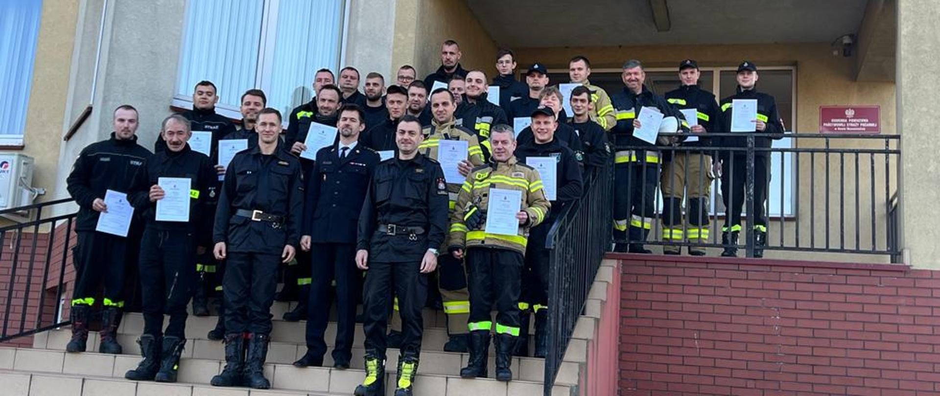 Na schodach zewnętrznych, prowadzących do drzwi wejściowych do komendy, kolejno na stopniach ku górze stoją druhowie OSP, którzy ukończyli szkolenie podstawowe, ubrani w umundurowanie koszarowe, trzymający w dłoniach zaświadczenia o ukończeniu szkolenia. Na samym dole, w pierwszym rzędzie stoi Zastępca Komendanta Powiatowego oraz komisja egzaminacyjna, składająca się z oficerów Komendy Powiatowej Straży Pożarnej w Rawie Mazowieckiej.