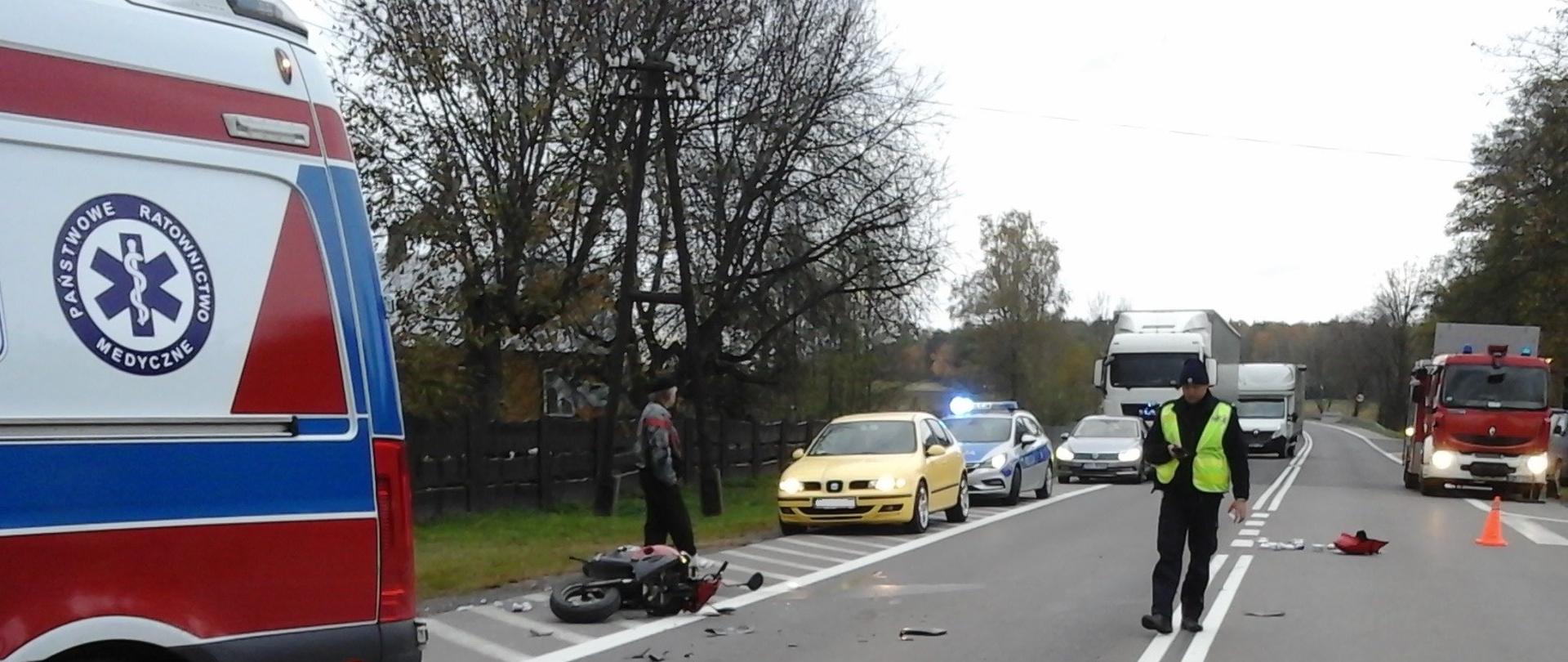 Leżący na jedni uszkodzony skuter. Obok karetka pogotowia ratunkowego. W tle radiowóz, samochód ratowniczo gaśniczy i pojazdy stojące w korku.