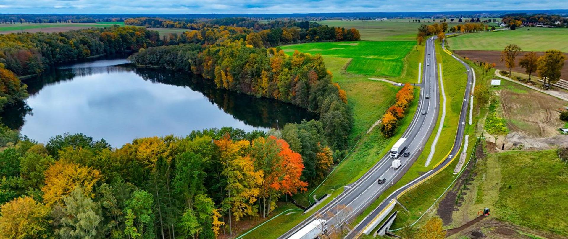 Widok z lotu ptaka na drogę krajową biegnącą wzdłuż jeziora otoczonego lasem w jesiennych barwach. Po prawej stronie widać pola uprawne, a po lewej gęsty las z drzewami w odcieniach zieleni, żółci i pomarańczu. Na drodze poruszają się samochody osobowe i ciężarowe, a w tle rozciąga się pagórkowaty krajobraz.