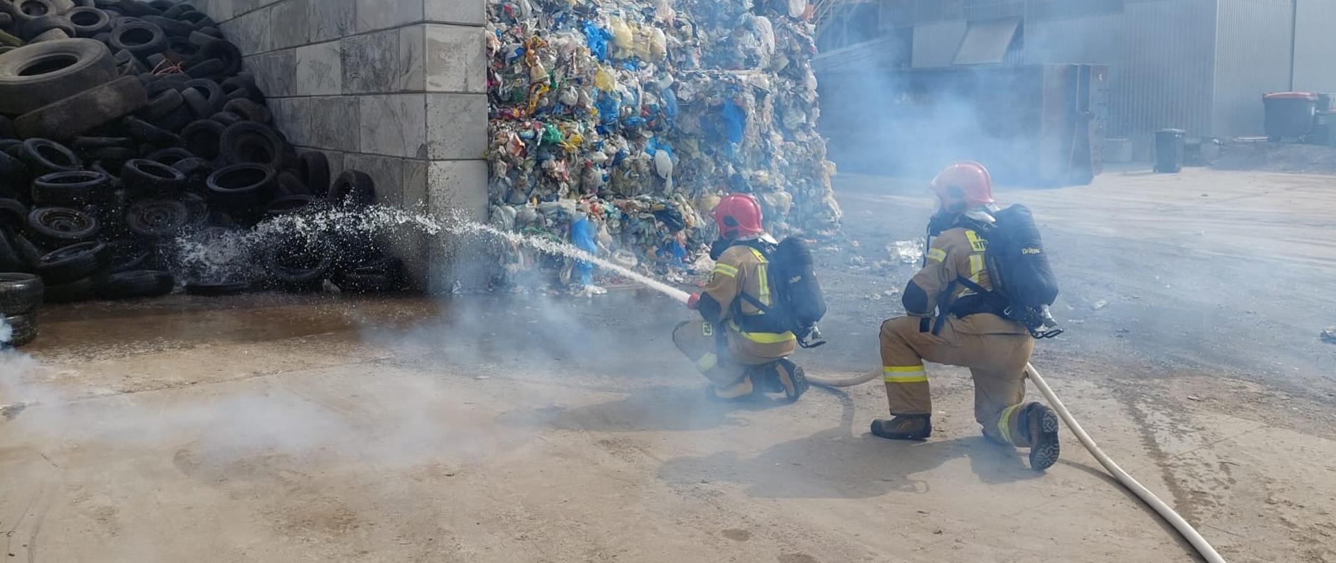 Dwóch strażaków ubranych w aparaty ochrony układu oddechowego podaje prąd wody na opony. Widać zadymienie.