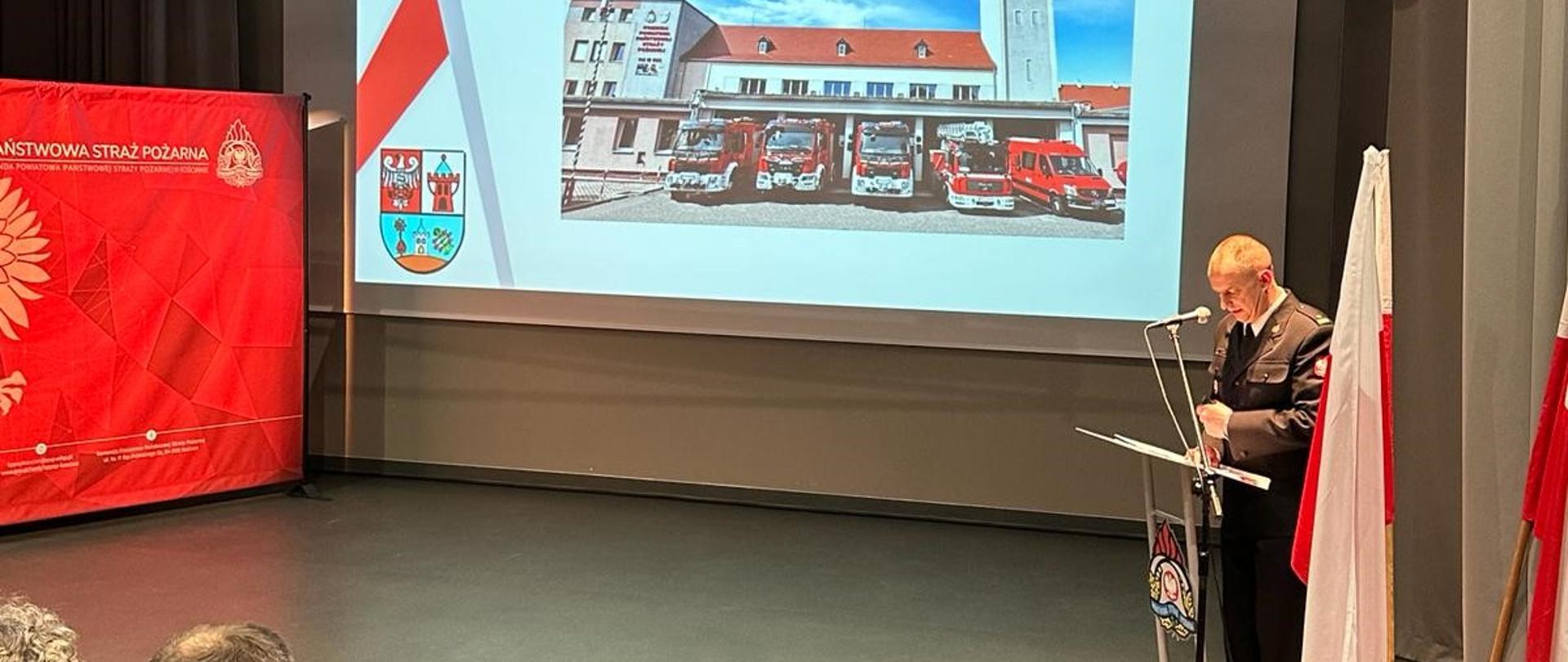 Kościańscy strażacy podsumowali 2023 rok. Strażacy i zaproszeni goście zebrani na sali podczas narady rocznej. Funkcjonariusz przemawia, w tle ekran z prezentowanym materiałem.