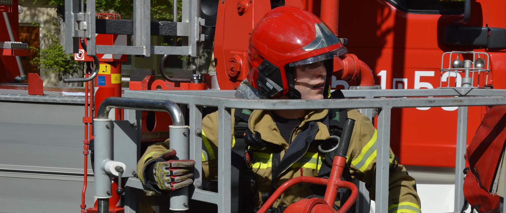 Ćwiczenia w DPS. na pierwszym planie kosz specjalnego pojazdu pożarniczego z podnośnikiem hydraulicznym. W podnośniku znajduje sie strażak w czerwonym hełmie. W tle fragment budynku i zieleni.