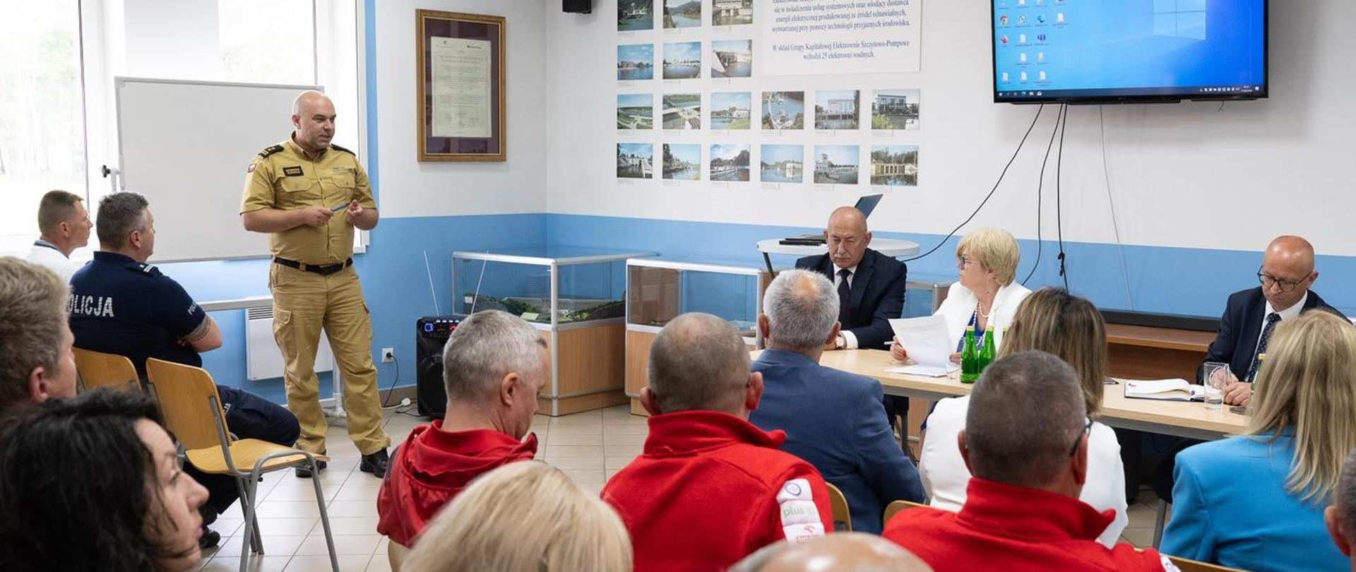 Zdjęcie zrobione w sali szkoleniowej, podczas narady Wojewódzkiego Zespołu Zarządzania Kryzysowego. Na fotografii oficer Państwowej Straży Pożarnej w stopniu starszego brygadiera prowadzi wykład. Na sali siedzą uczestnicy spotkania.