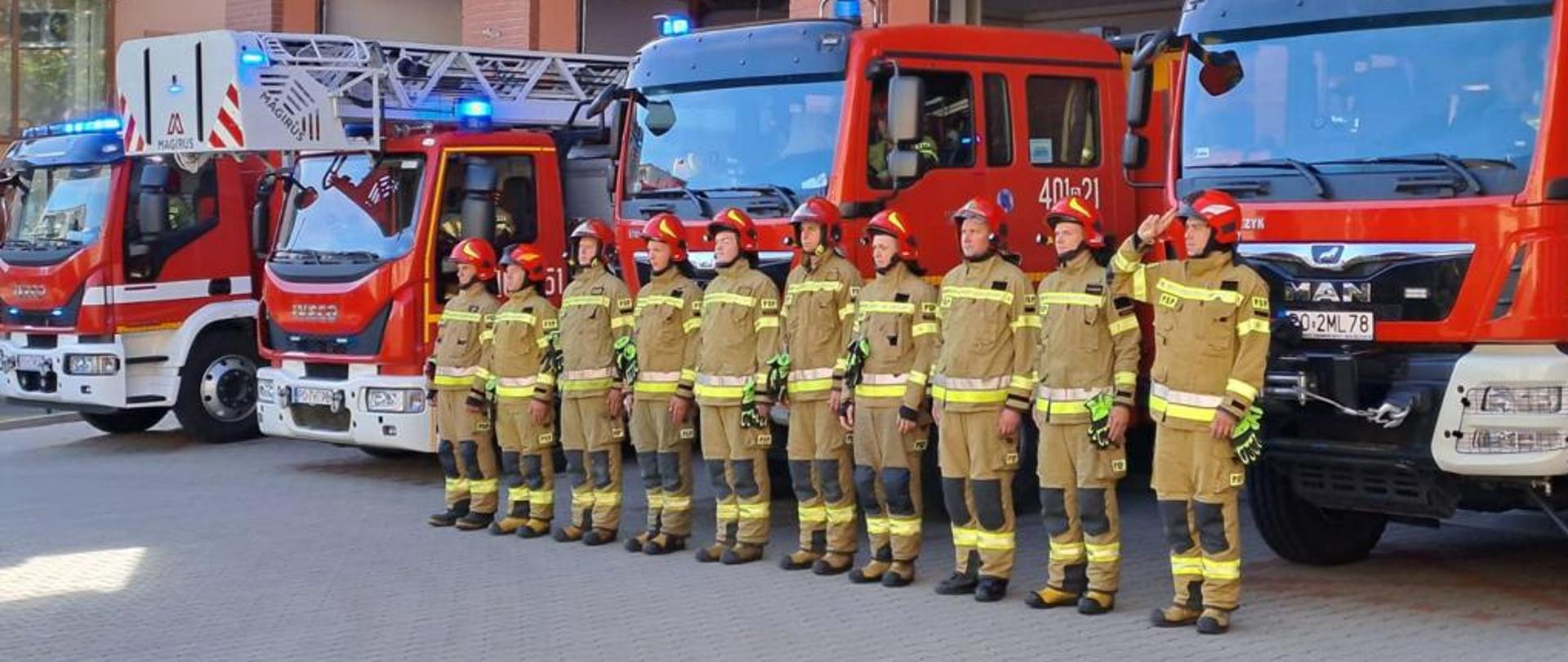 Strażacy stoją w szeregu, w pozycji "baczność", w tle samochody strażackie i budynek komendy