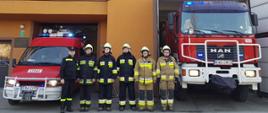 Widok z przodu. Sześciu strażaków OSP Brzeźnica stoi w ubraniach specjalnych przed samochodami pożarniczymi przed garażem OSP. 