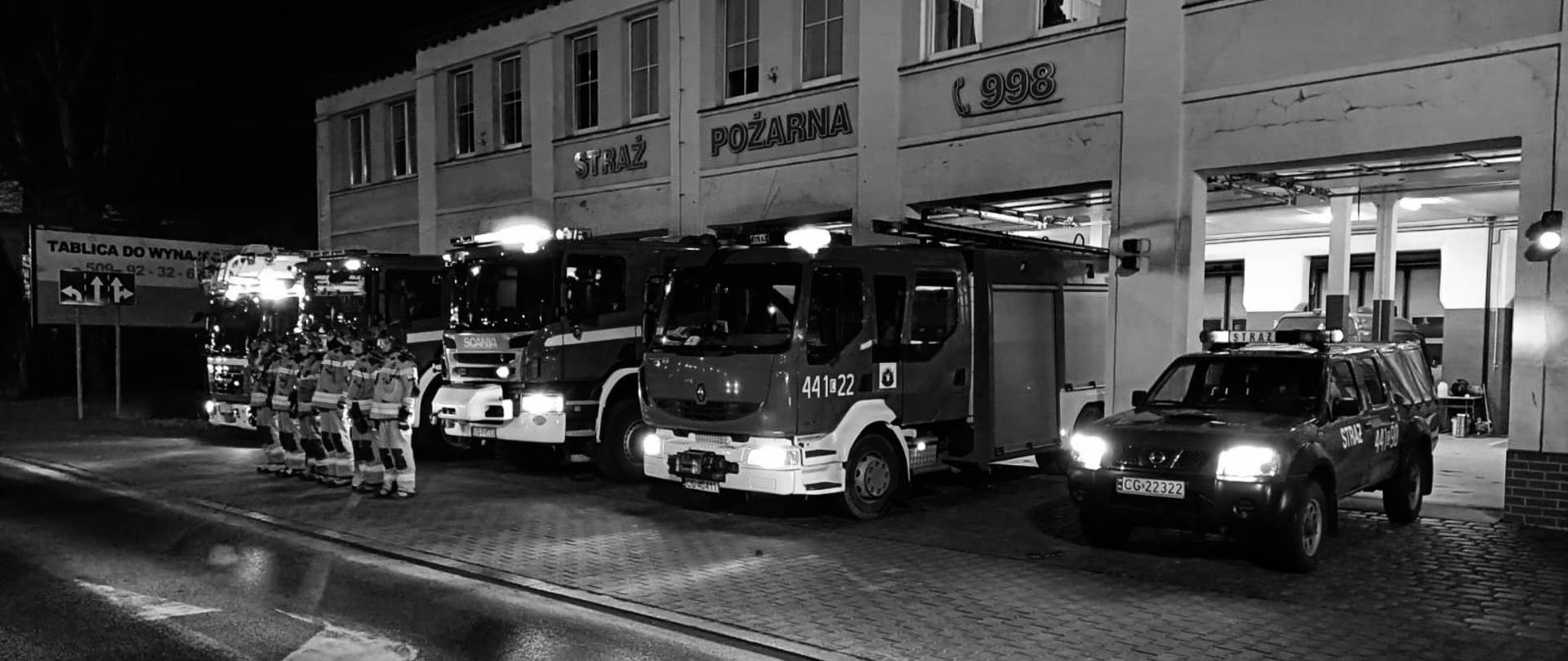 Czarno-białe zdjęcie przedstawia budynek z napisem straż pożarna 998 po zmierzchu. Przed otwartymi bramami garażowymi stoi 5 wozów strażackich z włączonymi światłami mijania i światłami alarmowymi. Przed pojazdami w szeregu stoją strażacy w ubraniach bojowych i hełmach. Jeden z nich oddaje honor