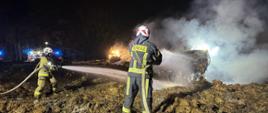 Nocna akcja gaśnicza na składowisku słomy. Czterech strażaków w pełnym umundurowaniu ochronnym i hełmach operuje wężami strażackimi, kierując strumienie wody na dymiące pryzmy materiału organicznego. W tle widoczny jest pracujący sprzęt ciężki (ładowarka) oraz łuna światła przebijająca się przez kłęby białego dymu.